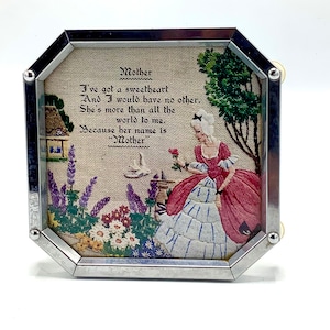 Könnte beinhalten: Eine gerahmte Stickerei mit einem Gedicht über eine Mutter. Die Stickerei zeigt eine Frau in einem blau-weißen Kleid, die eine rosa Rose in einem Garten hält. Das Gedicht lautet: "Mutter Ich habe einen Schatz Und ich möchte keinen anderen haben. Sie ist mehr als die ganze Welt für mich. Denn ihr Name ist "Mutter" "