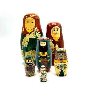 May include: A set of six hand-painted wooden nesting dolls, or matryoshka dolls, depicting a traditional nativity scene. The dolls are painted in bright colors and feature detailed designs. The largest doll is a woman in a green dress, holding a baby in her arms. The other dolls depict a man in a red robe, a man in a blue robe, a man in a brown robe, and a man in a white robe.