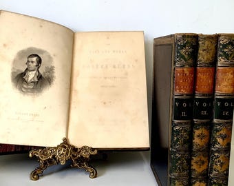 Pode incluir: Um conjunto de quatro livros antigos com capas em relevo dourado. Os livros são intitulados "The Works of Robert Burns" e são os volumes II, III e IV. Os livros estão sobre uma superfície branca.