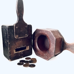 Könnte beinhalten: Zwei antike Holzmünzdosen, eine mit einem Scharnierdeckel und einem Schlitz für Münzen, die andere mit einer runden Öffnung und einem Griff. Beide sind dunkelbraun und haben eine abgenutzte Oberfläche. Ein Haufen alter Münzen liegt vor den Dosen.