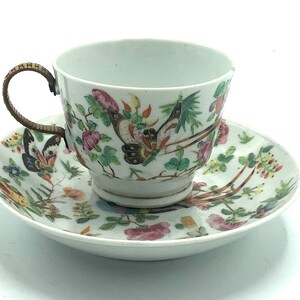 May include: A white porcelain teacup and saucer set with a floral and butterfly design. The cup has a gold rim and a brass handle. The saucer has a matching floral and butterfly design.
