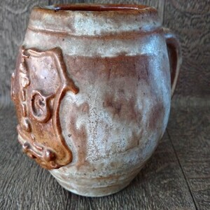 May include: A brown and white ceramic mug with a raised design on the front. The design features a stylized face with a crown and a beard.
