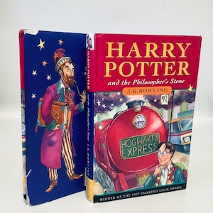Puede incluir: Un libro de tapa dura rojo con una ilustración azul y dorada de un tren en la portada. El título del libro es "Harry Potter y la piedra filosofal" de J.K. Rowling. El libro ganó el premio Smarties Gold Award en 1997.