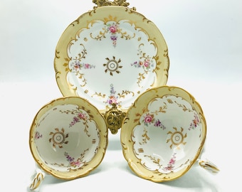 May include: A set of three vintage porcelain teacups and a saucer with a gold trim and floral pattern. The cups and saucer are decorated with delicate pink and purple flowers.