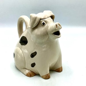 Puede incluir: Una tetera de cerámica con forma de cerdo con manchas marrones y un glaseado marrón claro. El cerdo tiene la boca abierta y ojos grandes. La tetera tiene un asa y un pico.