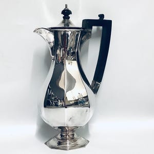 Op de afbeelding: Een zilveren theepot met een zwarte handgreep en een zilveren deksel. De theepot heeft een rond lichaam met een geribbeld ontwerp en een ronde basis.