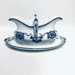 Könnte beinhalten: Eine weiße Porzellansauce mit einem blauen Blumendesign und einer passenden Untertasse. Die Sauciere hat einen Griff und einen Ausguss.