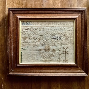 May include: A framed cross-stitch embroidery with a brown wooden frame. The embroidery features a floral design with the letters "ABC" and the number "456" visible.