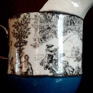 May include: A white ceramic pitcher with a blue base and a black and white print of a pastoral scene. The print features a tree, a river, and people fishing.