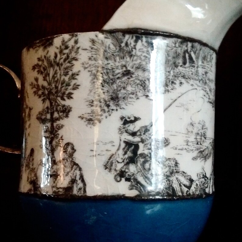 May include: A white ceramic pitcher with a blue base and a black and white print of a pastoral scene. The print features a tree, a river, and people fishing.