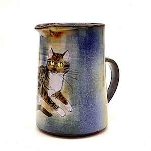 Puede incluir: Una jarra de cerámica marrón y blanca con un esmalte azul y una imagen pintada a mano de un gato atigrado con ojos amarillos.
