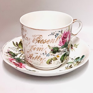 May include: A white teacup and saucer with a floral design and gold lettering that reads "To Present From/ Bully".