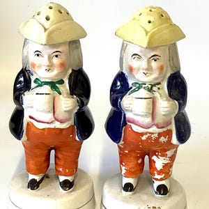 May include: Pair of vintage salt and pepper shakers shaped like men wearing colonial hats and holding cups. The shakers are painted in blue, orange, white, and green.
