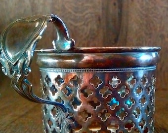 May include: A silver-plated cup holder with an ornate handle and a decorative, openwork design. The holder is made of metal and has a shiny, polished finish.