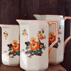 May include: Three white ceramic pitchers with floral designs. The pitchers have a red, orange, yellow, blue, and green floral pattern. The pitchers are all different sizes.