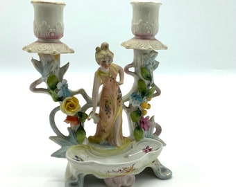 May include: A vintage porcelain double candlestick holder featuring a woman figurine and floral accents. The candlestick holders are white with pink trim, and the base is decorated with colorful flowers and leaves. A decorative piece for a table or shelf.