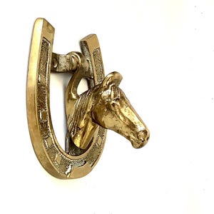 May include: A gold-toned brass door knocker shaped like a horseshoe with a horse's head protruding from the top. The horseshoe has a detailed design.