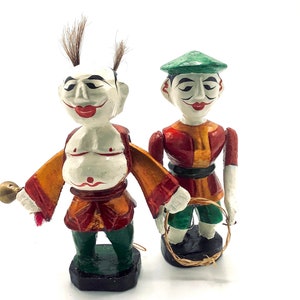 Puede incluir: Dos figuritas de madera con caras pintadas y ropa colorida. La figurita de la izquierda sostiene una pequeña campana y la figurita de la derecha sostiene un aro.