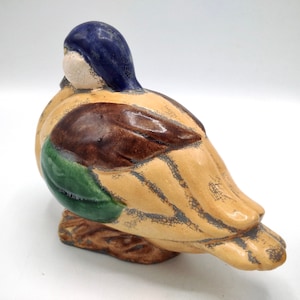 Pu&ograve; includere: Una statuetta di anatra in ceramica con testa blu, ali marroni e verdi e corpo beige screpolato.
