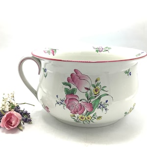 Puede incluir: Bote de cerámica blanca con un diseño floral con rosas rosas, flores amarillas y hojas verdes. El bote tiene un borde rojo y una sola asa.