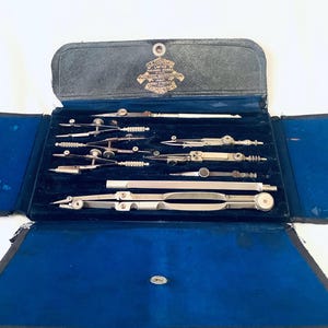 May include: A vintage set of drafting instruments in an open case. The case is black with a blue interior, housing various silver-coloured compasses and dividers. A metal emblem is visible on the case lid.