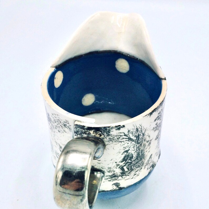 May include: A white ceramic creamer with a blue interior and white polka dots. The creamer has a silver handle and a black and white printed design on the exterior.