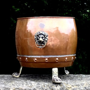 May include: A copper planter with a lion head design and three decorative feet. The planter is sitting on a stone surface.
