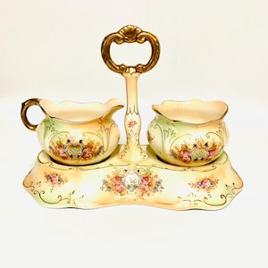 Puede incluir: Juego de azucarero y lechera de porcelana color crema con diseños florales. El juego incluye una lechera con asa dorada y un azucarero, ambos sobre una bandeja a juego con asa dorada. El juego está decorado con flores coloridas.