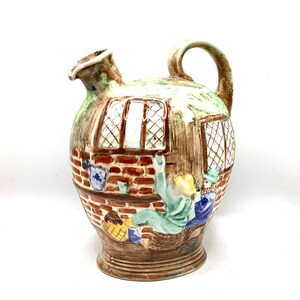 May include: A brown ceramic pitcher with a scene of people in a brick building. The pitcher has a green glaze and a handle.