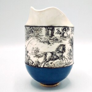 May include: A white ceramic pitcher with a blue base and a black and white print of a horse and rider. The print is a repeating pattern of a horse and rider in a field.