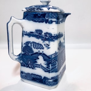 May include: A blue and white ceramic pitcher with a lid. The pitcher features a classic Willow pattern design with a handle and a spout. The pitcher is rectangular with rounded corners and measures approximately 8 inches tall.