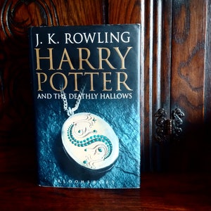 May include: A hardcover book titled "Harry Potter and the Deathly Hallows" by J.K. Rowling. The cover features a silver locket with green gemstones on a dark blue background.