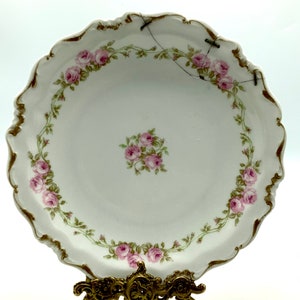 May include: A white porcelain plate with a gold rim and a floral design of pink roses. The plate is on a gold metal stand.