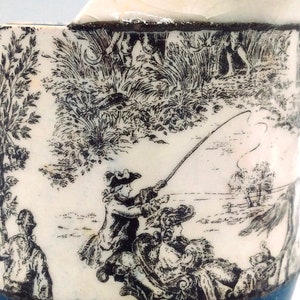 May include: A white ceramic mug with a blue rim and a silver handle. The mug features a black and white print of a fishing scene with a man in a hat casting a line.