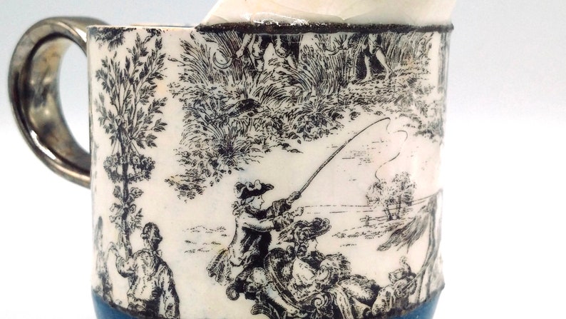 May include: A white ceramic mug with a blue rim and a silver handle. The mug features a black and white print of a fishing scene with a man in a hat casting a line.