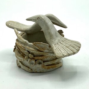 May include: A ceramic sculpture of two birds in a nest. The birds are white with brown accents and have a stylized, whimsical look. The nest is made of brown and white clay and has a textured surface.
