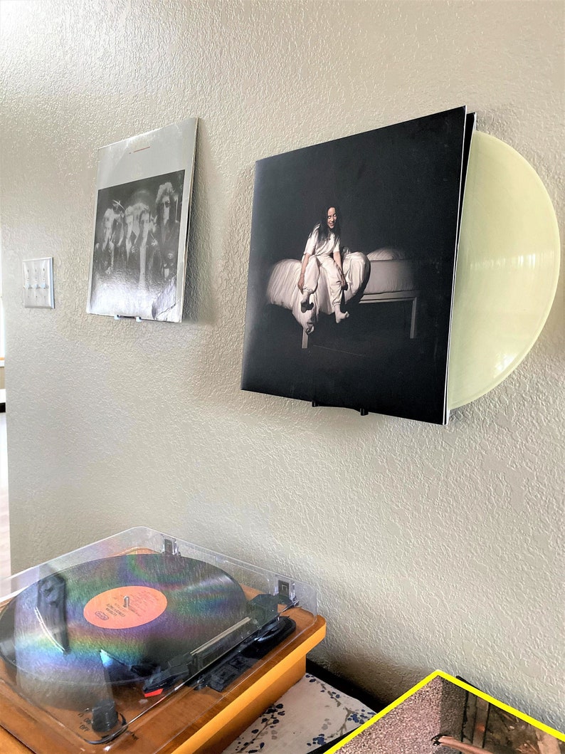 Puede incluir: Tres discos de vinilo expuestos en una pared, uno con un disco de vinilo transparente, otro con una imagen en blanco y negro de una mujer sentada en una cama y el &uacute;ltimo con una imagen en blanco y negro de un grupo de personas. Un tocadiscos con un disco encima est&aacute; sobre una superficie de madera debajo de los discos.