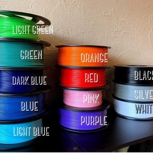 Puede incluir: Carretes de filamento en varios colores, incluyendo verde claro, verde, azul oscuro, azul, azul claro, naranja, rojo, rosa, morado, negro, plata y blanco. Los carretes est&aacute;n dispuestos en dos filas, con la fila superior que contiene los colores m&aacute;s claros y la fila inferior que contiene los colores m&aacute;s oscuros.