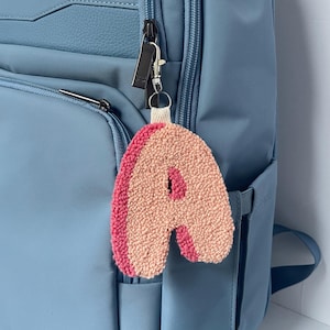 May include: A punch needle letter A keychain featuring a textured peach-colored front and a vibrant pink side border. The letter is attached to a silver-toned metal lobster clasp and a small cream-colored fabric loop, hanging from a blue backpack.