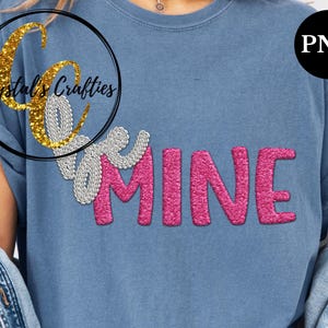 May include: A blue t-shirt with the words "Be Mine" in a sparkly design. The word "Be" is in silver, and "MINE" is in pink. The shirt also features a logo in a black circle with gold glitter text that says "Crystal's Crafties".