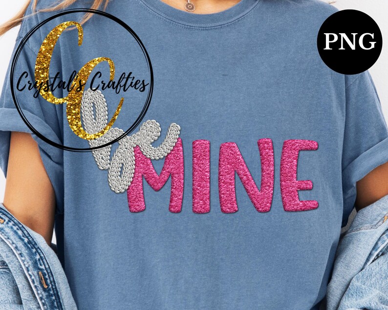 May include: A blue t-shirt with the words "Be Mine" in a sparkly design. The word "Be" is in silver, and "MINE" is in pink. The shirt also features a logo in a black circle with gold glitter text that says "Crystal's Crafties".