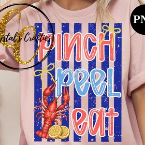 May include: Pink t-shirt with a graphic design featuring the words "Pinch Peel Eat" in red and blue, with a red crawfish and lemon slices. The design is set against blue and white vertical stripes.