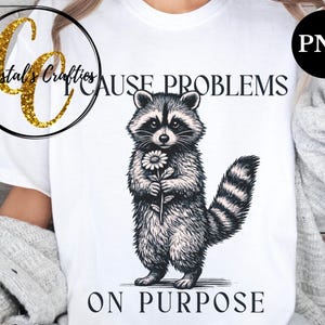 May include: White t-shirt featuring a detailed illustration of a raccoon holding a flower. The raccoon is in black and white, with the text "CAUSE PROBLEMS ON PURPOSE" in black. A gold glitter logo is in the upper left corner.