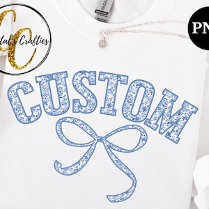 Puede incluir: Camiseta blanca con la palabra "CUSTOM" en azul con un patrón floral azul. Un lazo floral azul está debajo de la palabra.