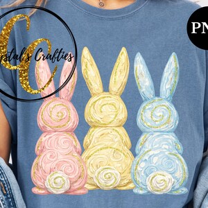 May include: A blue t-shirt featuring three Easter bunny designs in pink, yellow, and blue. Each bunny has a spiral design and gold glitter accents. The shirt also has the text "Crystal's Crafties" in a gold glitter font.