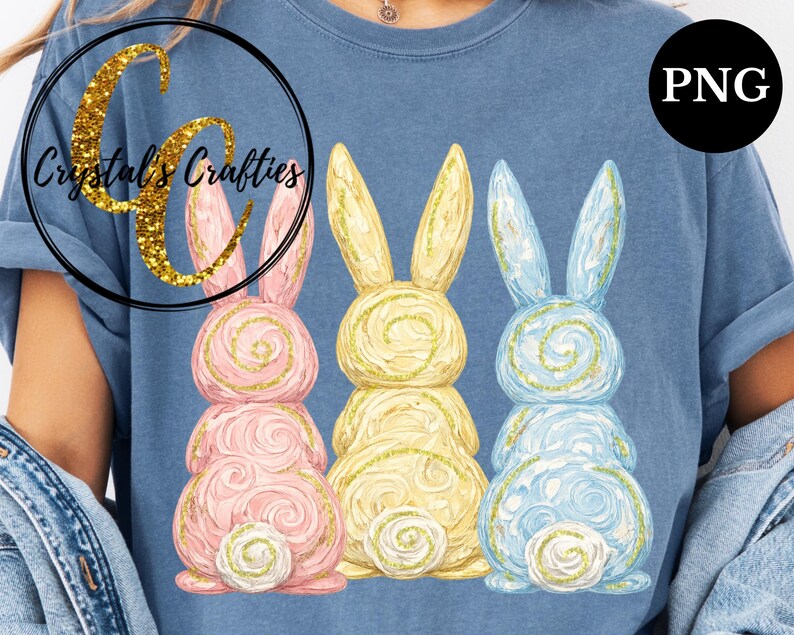May include: A blue t-shirt featuring three Easter bunny designs in pink, yellow, and blue. Each bunny has a spiral design and gold glitter accents. The shirt also has the text "Crystal's Crafties" in a gold glitter font.