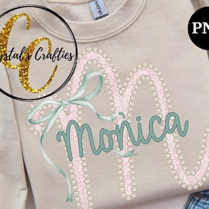 May include: A beige sweatshirt featuring a large, embellished letter "M" with the name "Monica" in teal script. The letter is outlined with pearls and pink lace, accented by a teal bow. The sweatshirt has the text "Crystal's Crafties" in a circle.