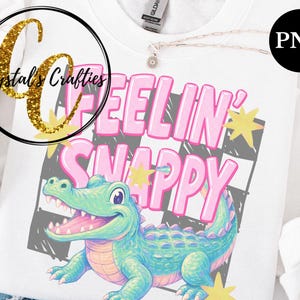 May include: White sweatshirt featuring a cartoon alligator with the text "FEELIN' SNAPPY" in pink letters. The alligator is light blue and green, with a pink mouth and a checkered background. The sweatshirt has a round neckline.