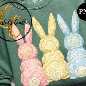 May include: A green sweatshirt featuring three Easter bunny designs in pink, yellow, and blue. Each bunny has a spiral design and gold glitter accents. The sweatshirt has the text "Crystal's Crafties" in a gold glitter font.