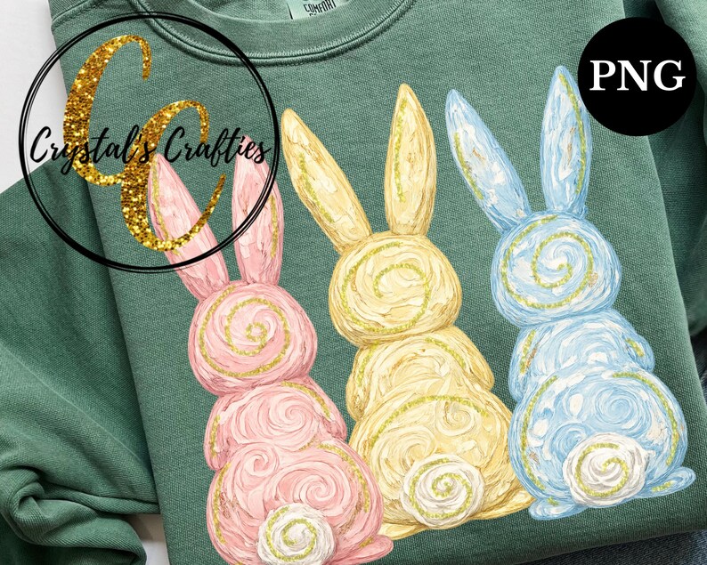 May include: A green sweatshirt featuring three Easter bunny designs in pink, yellow, and blue. Each bunny has a spiral design and gold glitter accents. The sweatshirt has the text "Crystal's Crafties" in a gold glitter font.